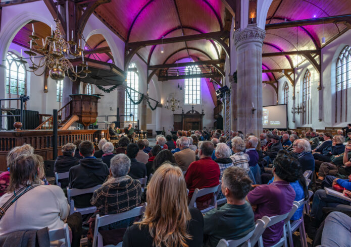 Kerkzaal, roze aangelicht, met honderden mensen op witte stoelen